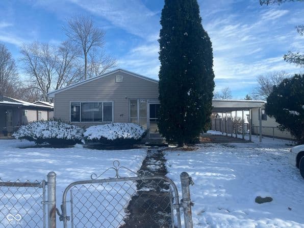 White residential home with snow-covered yard, tall evergreen tree, and chain-link fence